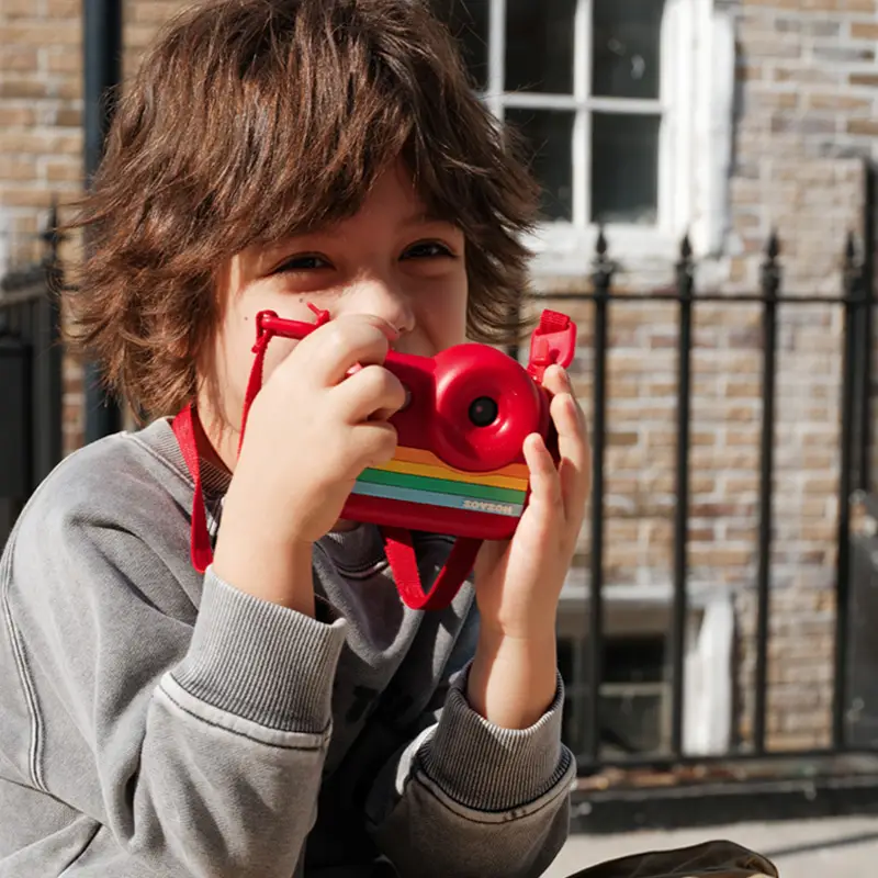 Uśmiechnięty chłopiec w szarej bluzie trzymający czerwony aparat fotograficzny Zoy Zoii przy oku