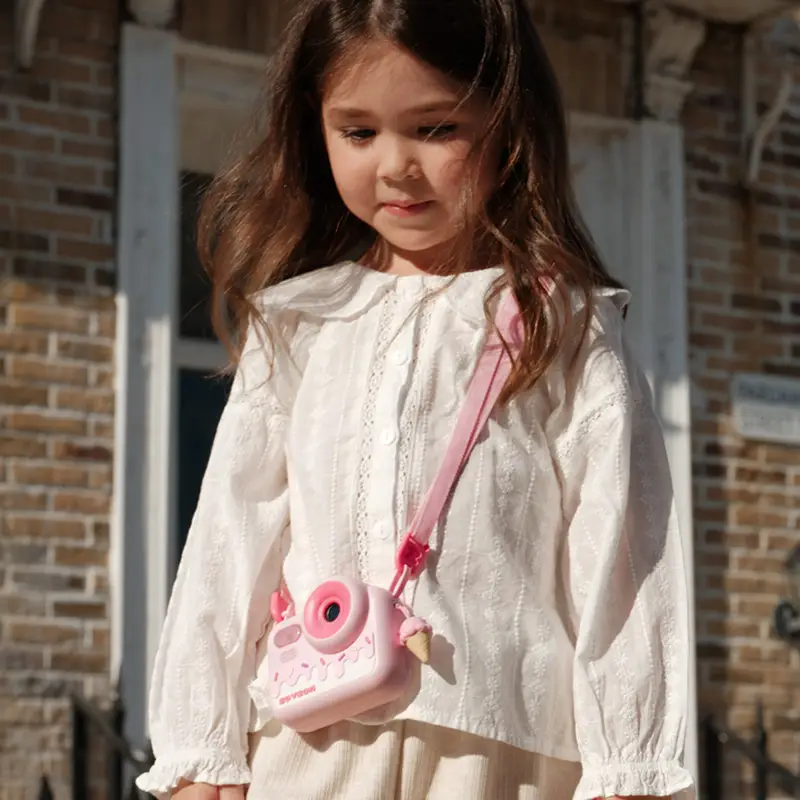 Uśmiechnięta dziewczynka w białej bluzce trzymająca różowy aparat fotograficzny na pasku